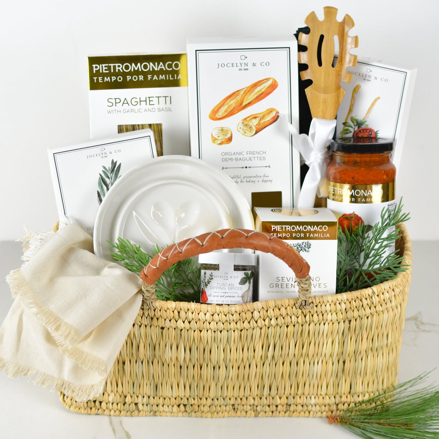 Gift basket featuring gourmet food items, including spaghetti, olive oil, French demi-baguettes, pasta sauce, dipping spices, and green olives. The basket includes a ceramic dish, wooden spaghetti server, and a neutral linen towel, all arranged in a woven basket with a leather-wrapped handle and festive greenery accents.