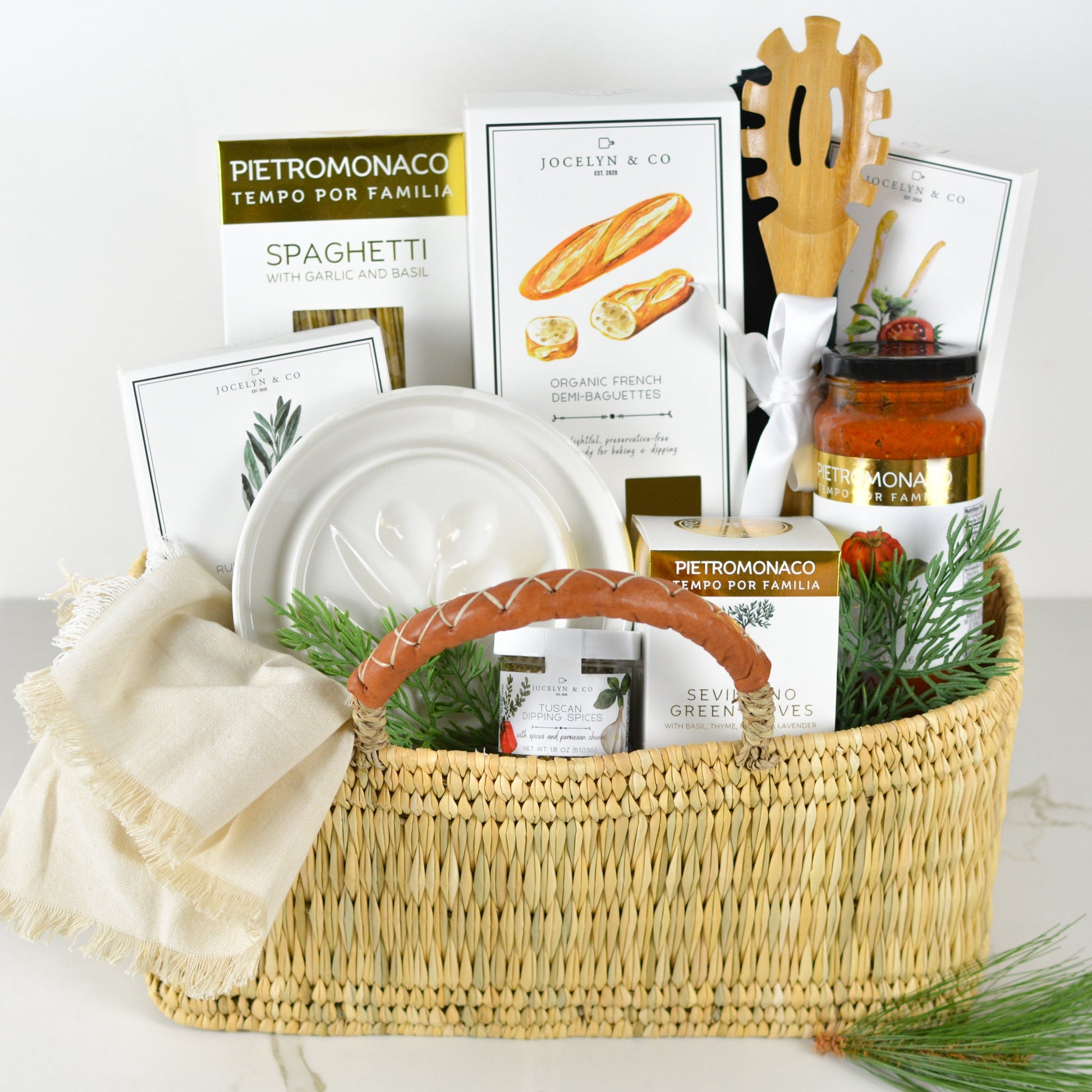 Gift basket featuring gourmet food items, including spaghetti, olive oil, French demi-baguettes, pasta sauce, dipping spices, and green olives. The basket includes a ceramic dish, wooden spaghetti server, and a neutral linen towel, all arranged in a woven basket with a leather-wrapped handle and festive greenery accents.