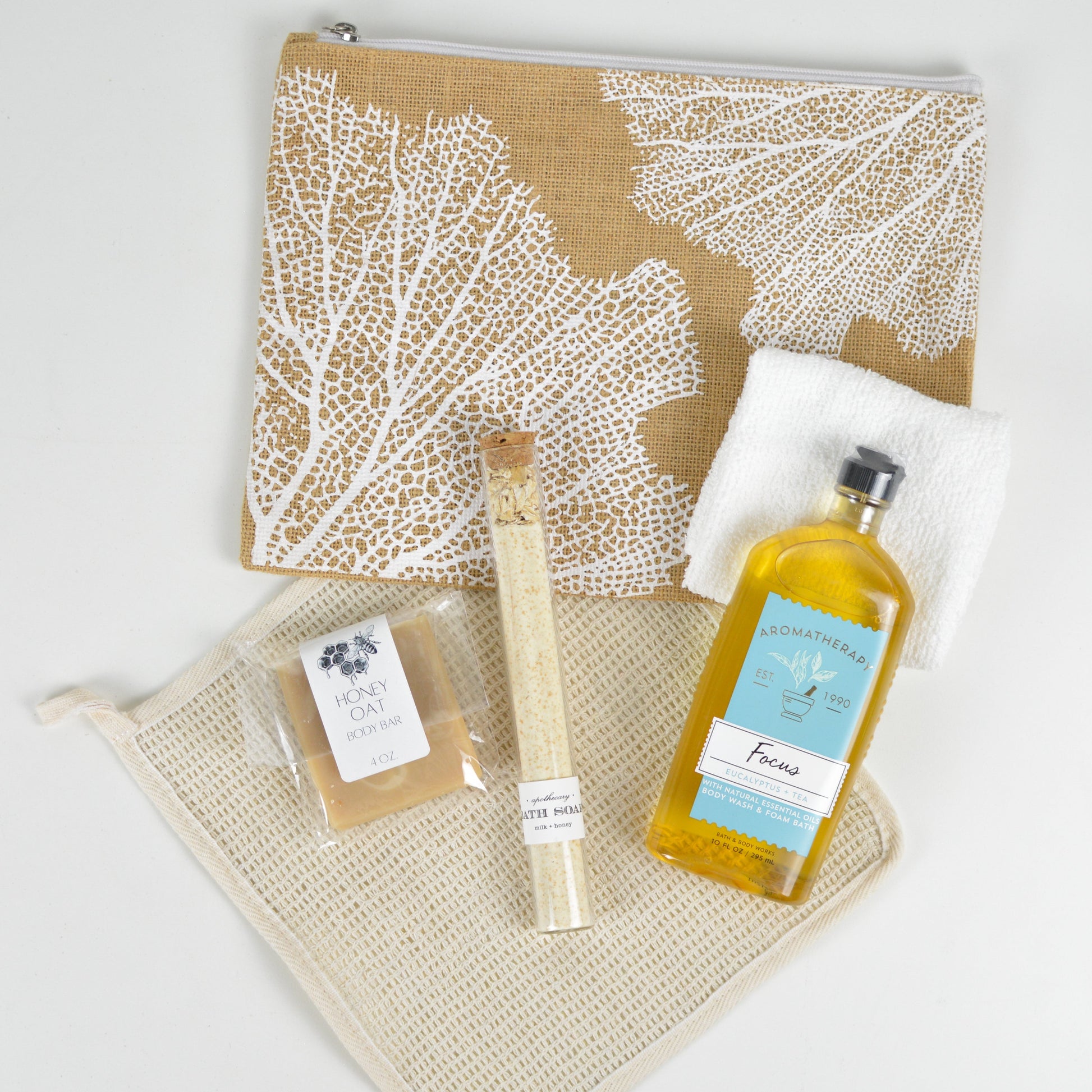 Printed jute makeup bag with sisal washcloth, white hand towel, honey oat body bar, tube of bath salts, and a bottle of Focus: Eucalyptus & Tea scented body wash