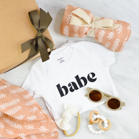 Kraft pinstripe gift box with baby sunglasses, wooden teether ring, white hair bow, muslin swaddling blanket, and white onesie with "babe" printed on front