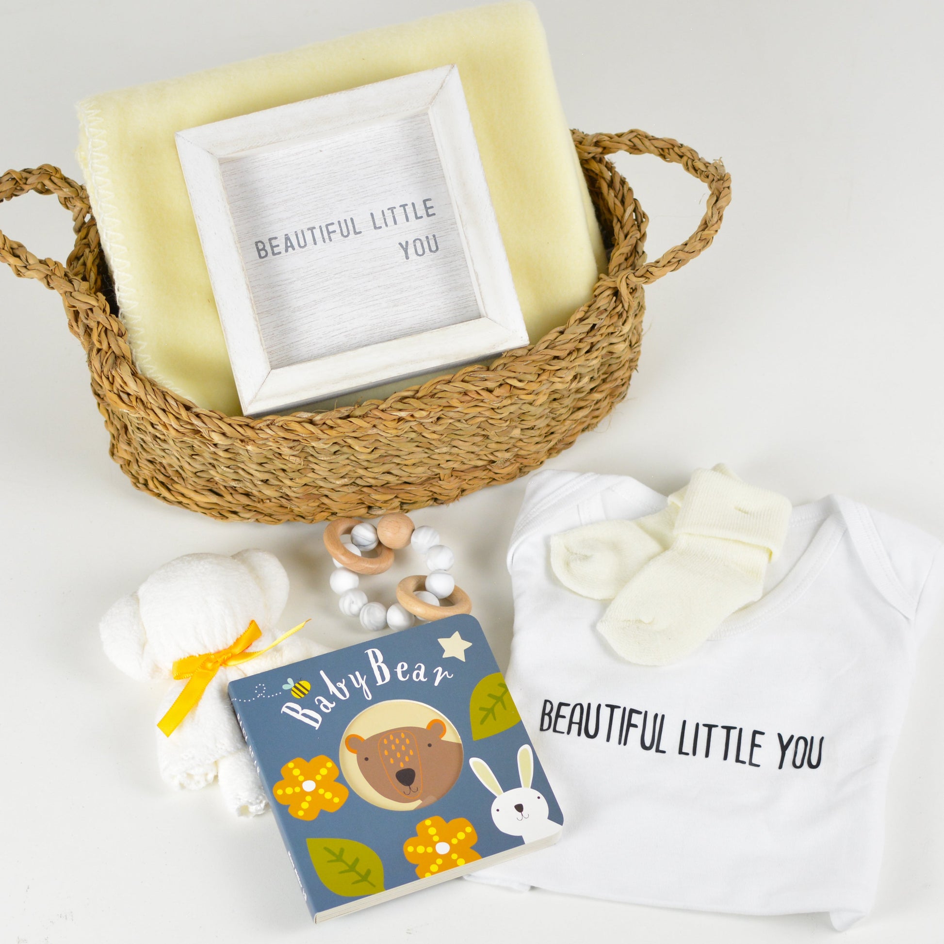 Seagrass basket with soft blanket, small wooden sign with saying "Beautiful Little You", onesie with saying "Beautiful Little You" printed on front, baby socks, plush bear, wooden teether ring, and Baby Bear board book