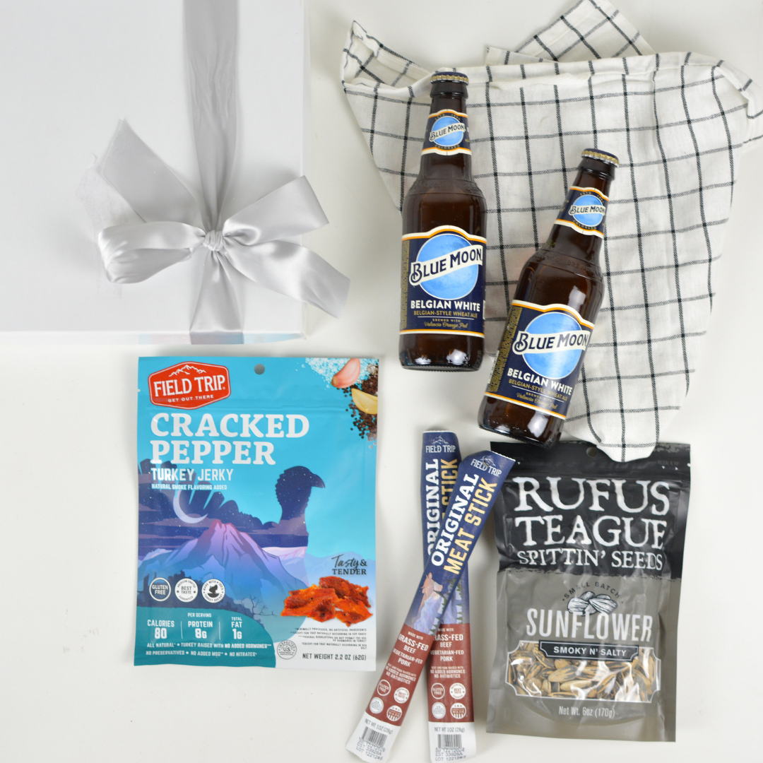 White gift box with bow, kitchen towel, two bottles of Blue Moon Belgian White Ale, package of turkey jerky, two meat stick snacks, and a package of sunflower seeds