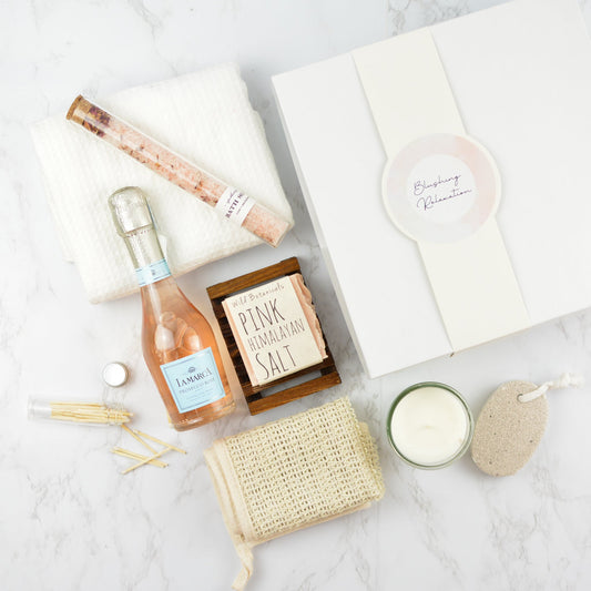 white gift box band that says "Blushing Relaxation", tube of bath salts, pumice stone, candle, bottle of match sticks, wood soap tray, sisal washcloth, white hand towel, pink himalayan salt bar of soap, and a small bottle of La Marca Prosecco Rose