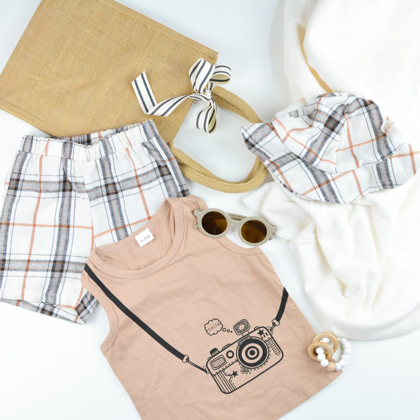 Jute gift bag with striped ribbon, plaid baby shorts and matching hat, baby sunglasses, wood teether ring,  sleeveless shirt with a print of a camera and "smile", and a plush white blanket