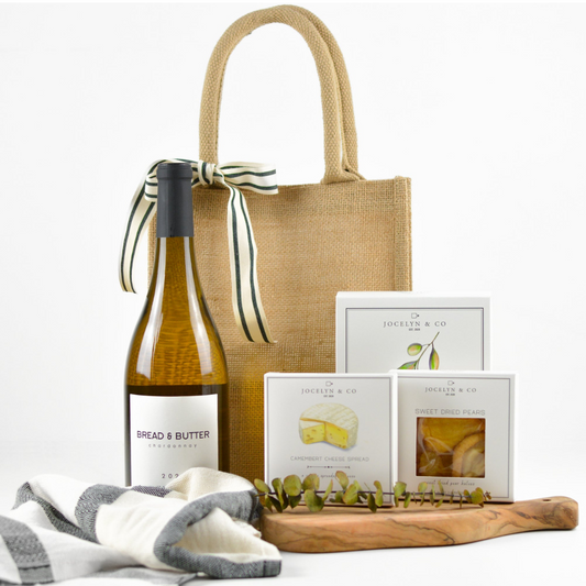 Jute tote with striped bow, Bread & Butter Chardonnay bottle, striped kitchen towel, cheese spread, dried pears, box of crackers, small cutting board 