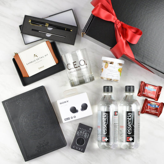 Black gift box with red bow,  gold and black pen, two Essentia water bottles, infinity cube fidget toy, black leather journal, earbud detail kit, jar of pistachios, Sony earbuds, two wrapped Ghirardelli chocolate squares, and cocktail glass printed with C.E.O. Consume Every Ounce 