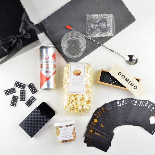 Black gift box with box,  slate board, two cocktail glasses, popcorn, box with dominos, black playing cards with metallic print, a jar of mixed nuts, Stoli Ginger Beer, and a bar spoon