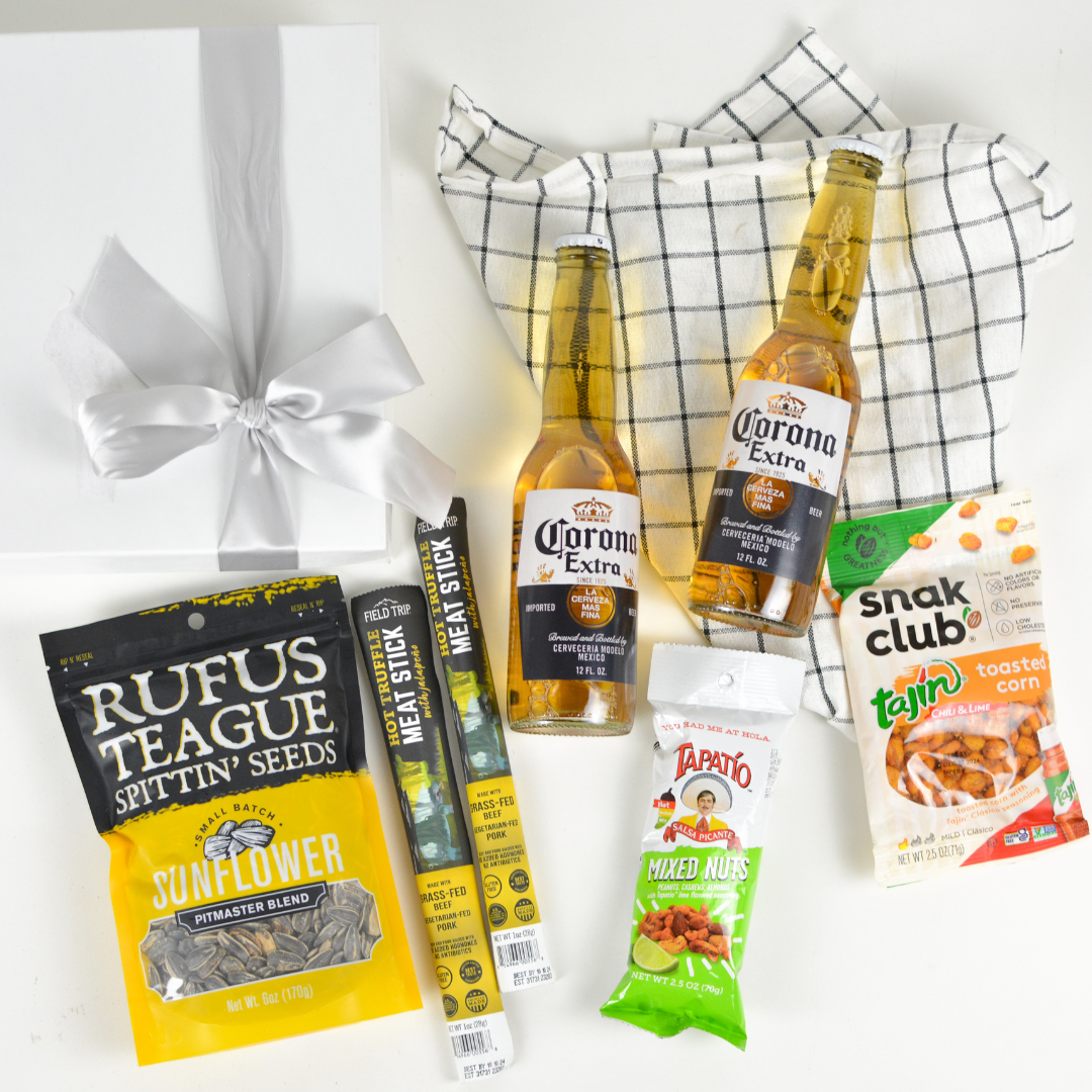 White gift box with bow, kitchen towel, two bottles of Corona Extra, package of sunflower seeds, two meat stick snacks, an individual size package of Tapatio mixed nuts, and an individual pack of Tajin toasted corn snacks