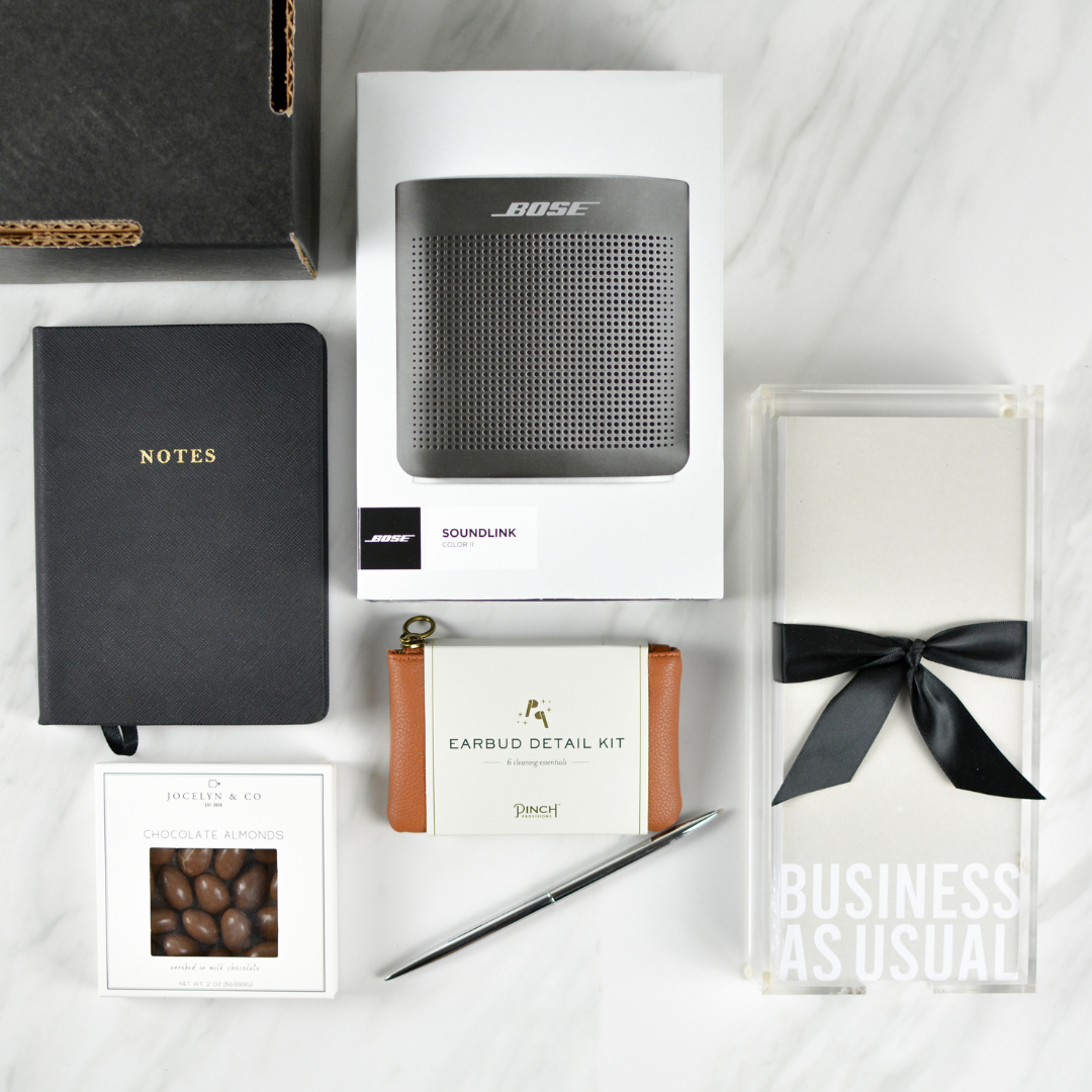 Black box with Bose speaker, "Business as Usual" notepad, black leather journal with NOTES in gold lettering, box of chocolate almonds, an earbud detail kit, and a pen