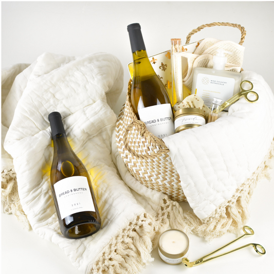 Basket with zig zag pattern, a quilted style white blanket with tassles, two bottles of Bread & Butter Chardonnay, a candle, a tube of match sticks, gold candle wick snippers, a sisal washcloth, a hair scrunchie, a gold and white dish with fleur-de-lei pattern, a bottle of hand lotion, and a tube of bath salts