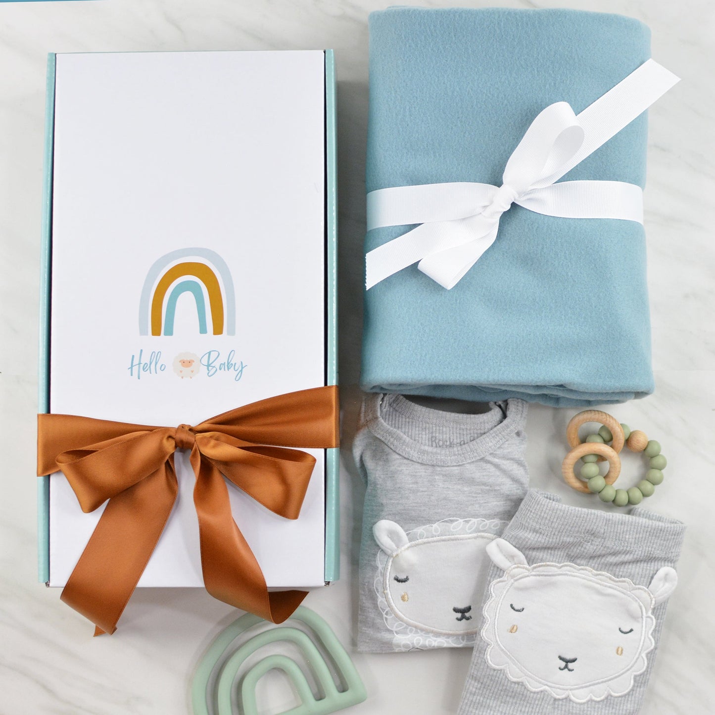 Gift box with rainbow, a sheep and "Hello Baby" printed on top, blanket, wood teether ring, rainbow silicone teeter, and a baby outfit with a lamb face on the front and backside of the pants and shirt.