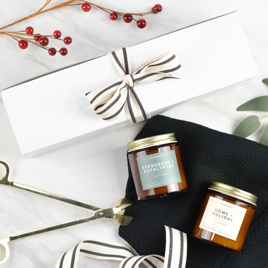 White gift box with stripe bow, two jar candles, a black kitchen towel, and a pair of candle wick trimmers