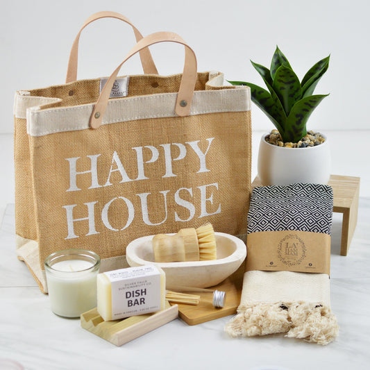 Jute tote with "Happy House" printed on outside, candle, jar of matches, dish soap bar, wooden soap dish, small dough bowl, small scrubber brush, kitchen towel with tassels, and plant