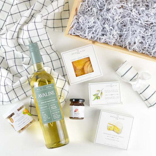 Wood tray with kitchen towel in a bow, bottle of organic white wine, box of dried pears, jar of pistachios, small jar of warm spiced apple jam, small box of crackers, and camembert cheese