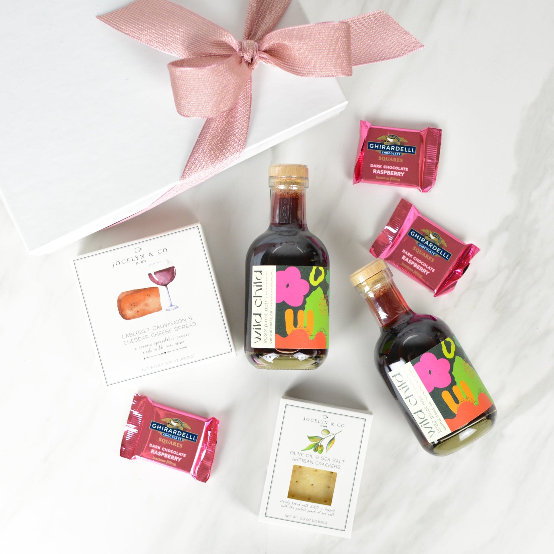 White gift box with pink box, cabernet cheese spread, small box of crackers, three pink Ghiradelli raspberry chocolate squares, and two small bottles of Wild Child pinot