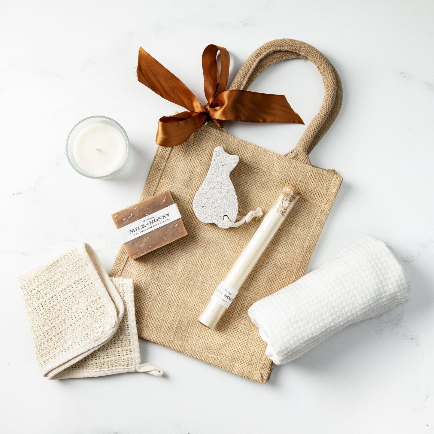 jute gift bag with white hand towel, sisal washcloth, candle, milk and honey soap bar, tube of bath salts, and pumice stone