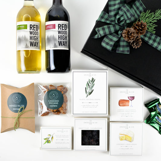 Black gift box with pine cones and bow, two bottles of wine, box of artisan crisps, candied pecans, box of mixed nuts, small box of crackers, box of dried cranberries, cabernet cheese spread, camembert cheese spread, two Ghirardelli mint chocolate squares