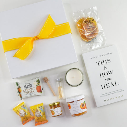 White gift box with bow, snack bar, two Tea Drops teas, candle, bottle of matches, mini jar of honey, jar of apricots, small package of crispy orange slices, and book, "When You're Ready This is How You Heal"